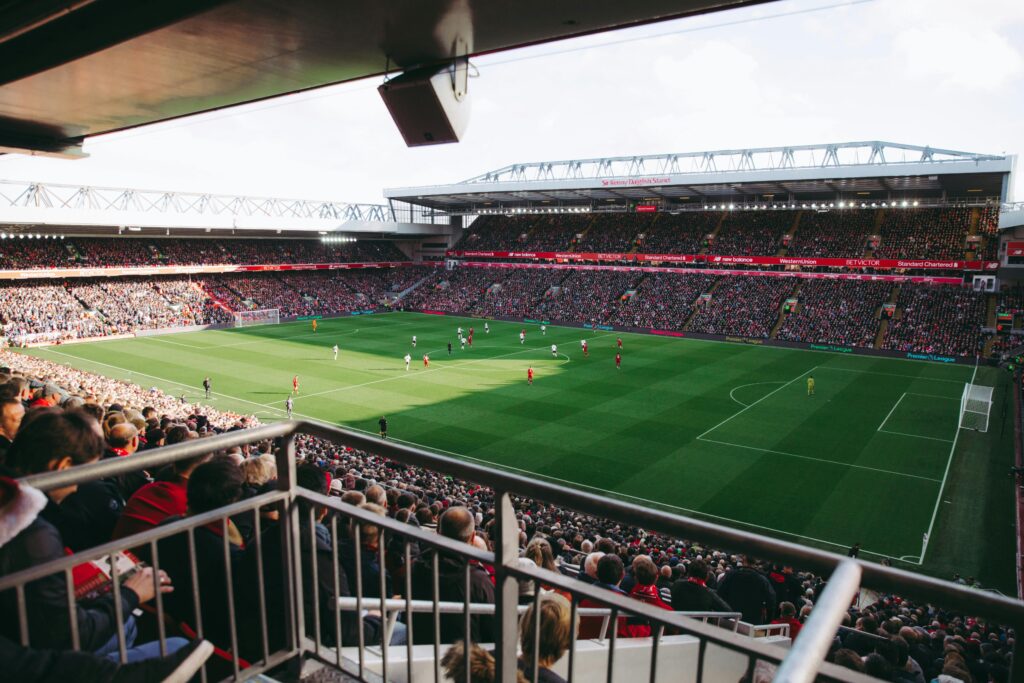 A packed Anfield stadium during a daytime Liverpool FC match, viewed from the stands behind supporters.