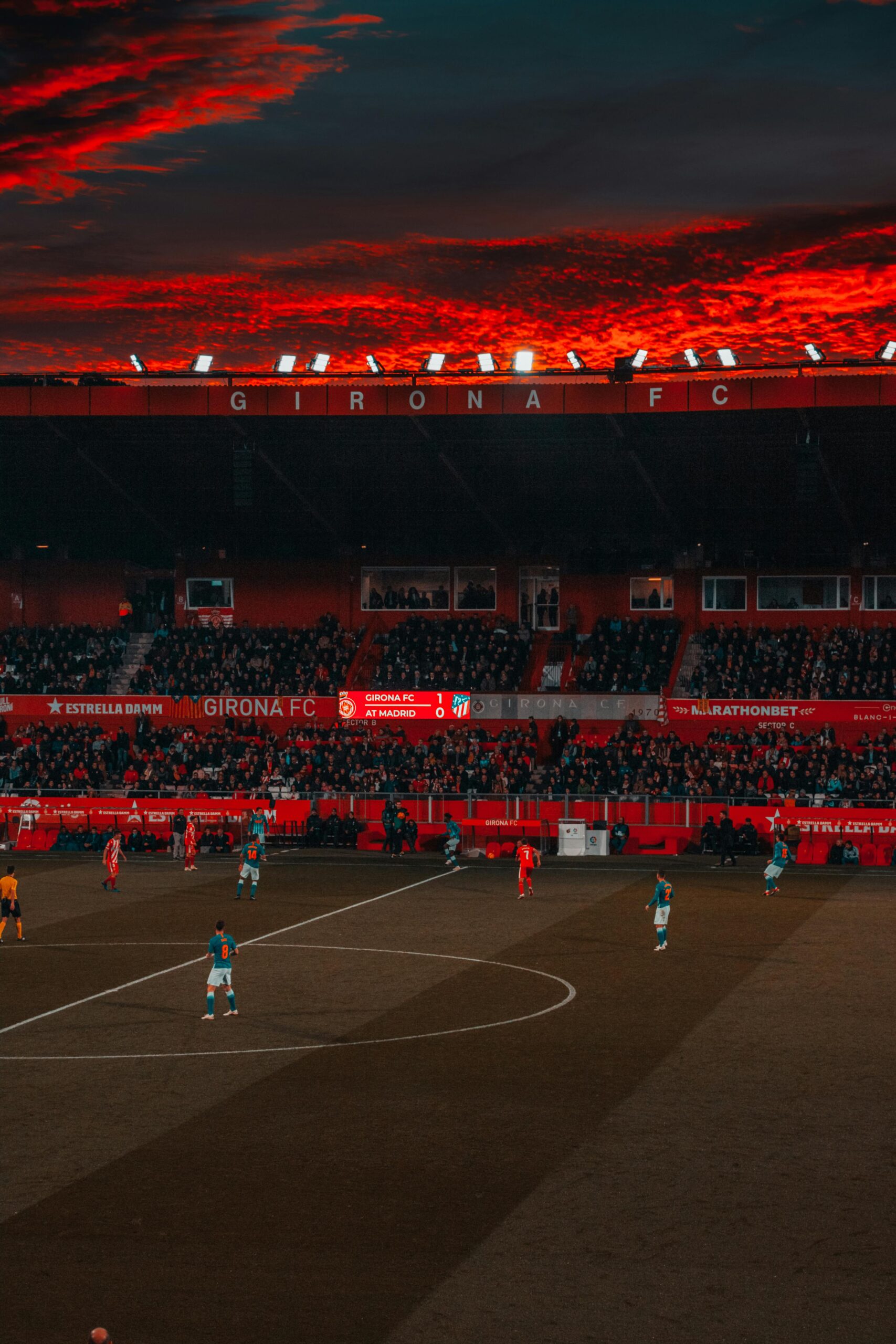 Evening football match at Girona FC’s stadium with a dramatic red sunset sky and supporters in the stands.