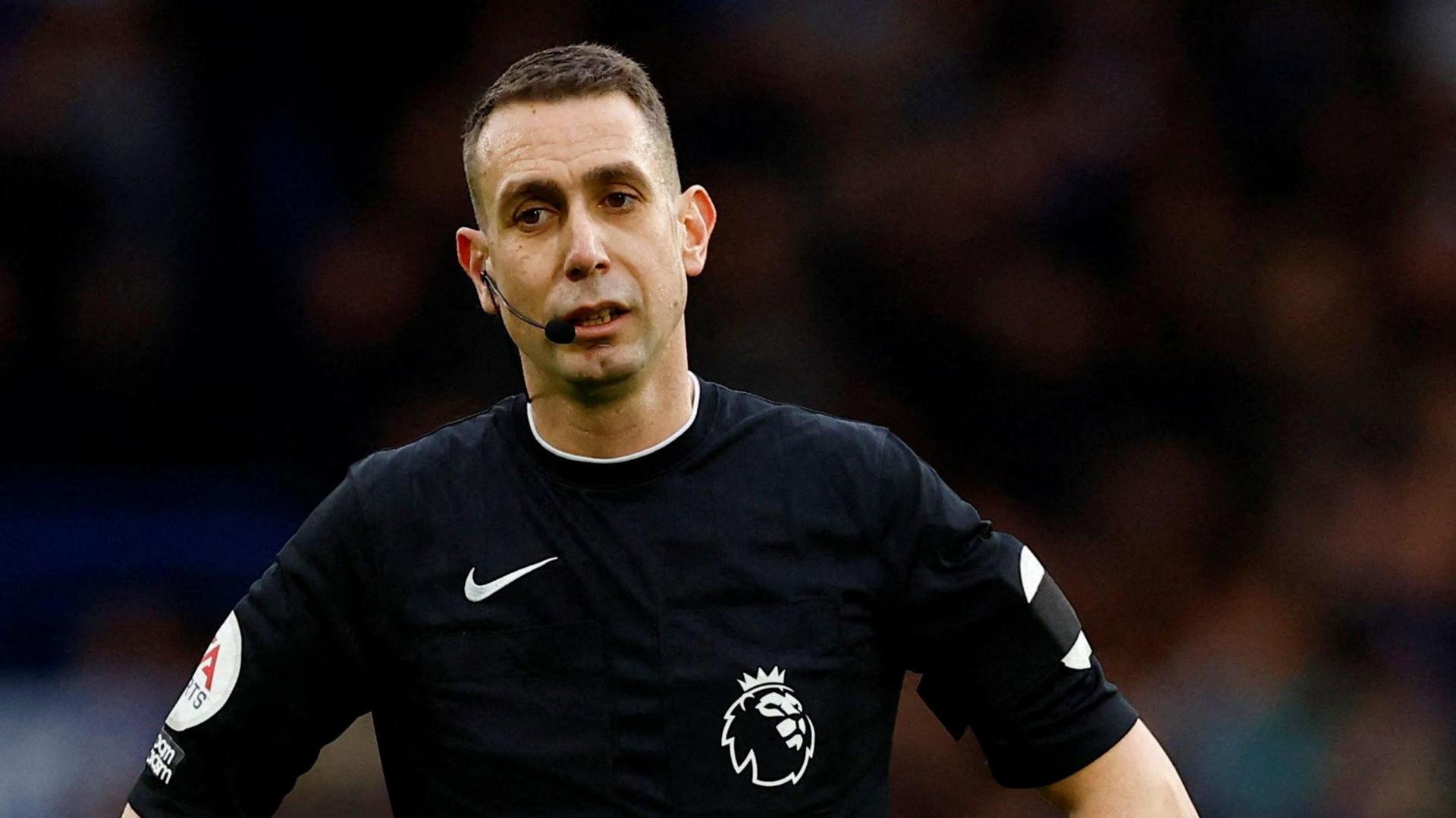 Premier League referee David Coote officiates a match while wearing the black officials kit and a communication headset.