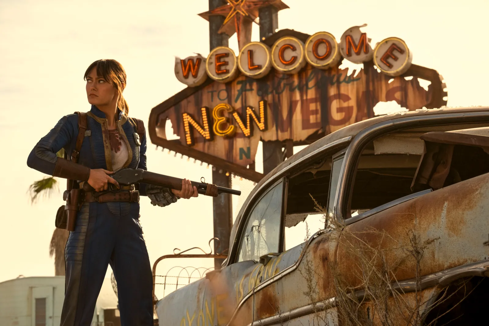 Lucy stands in front of the iconic New Vegas sign in the Fallout universe, wearing her Vault-Tec suit as neon lights glow behind her in the post-apocalyptic desert.