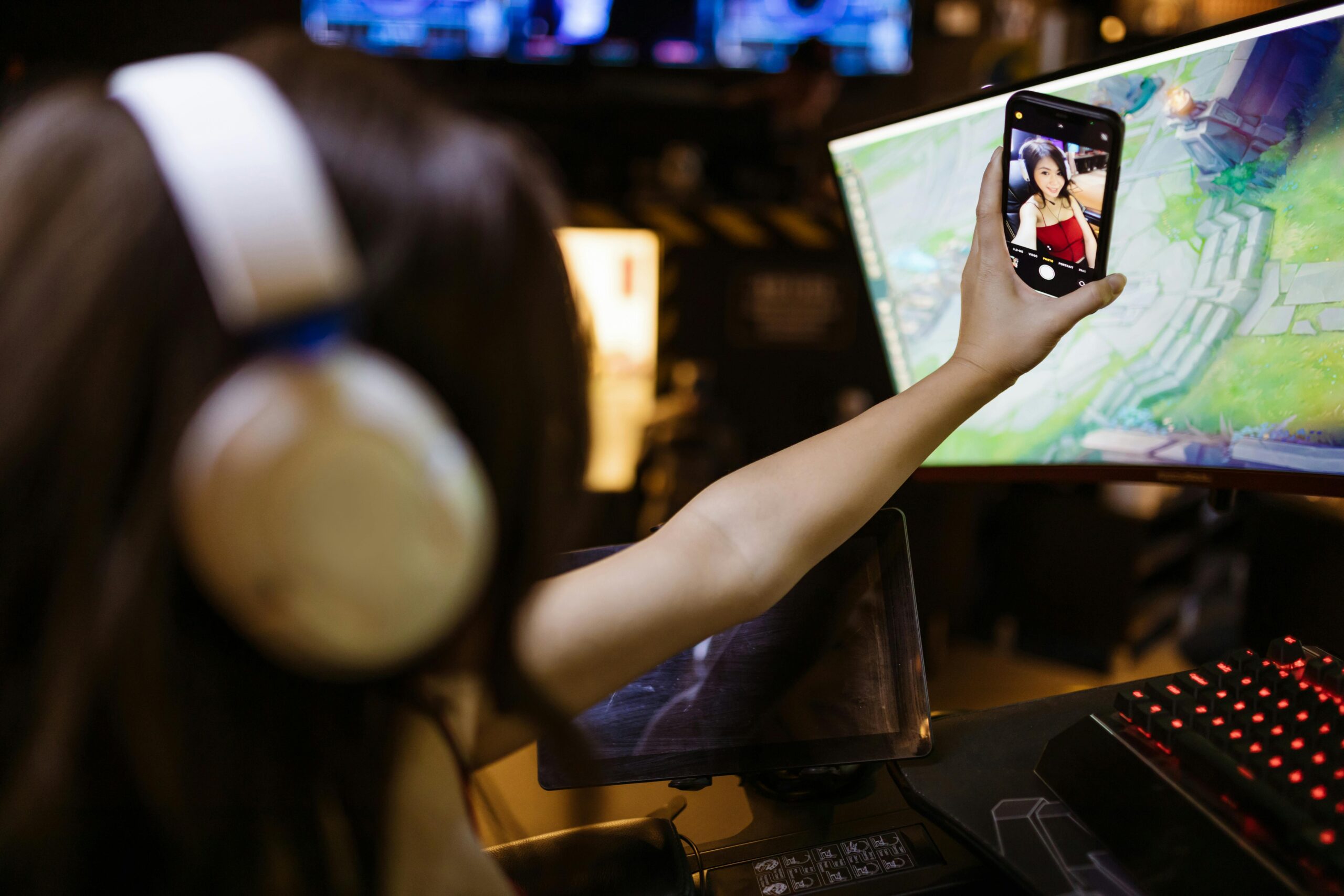 Female streamer taking a selfie during a gaming session with curved monitors and a headset.