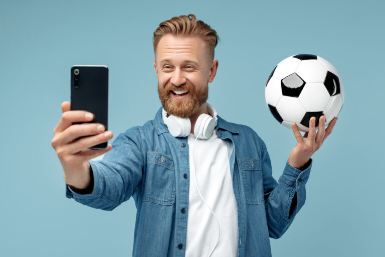 Smiling bearded hipster blonde young man take selfie on smartphone holding classic soccer ball over blue background